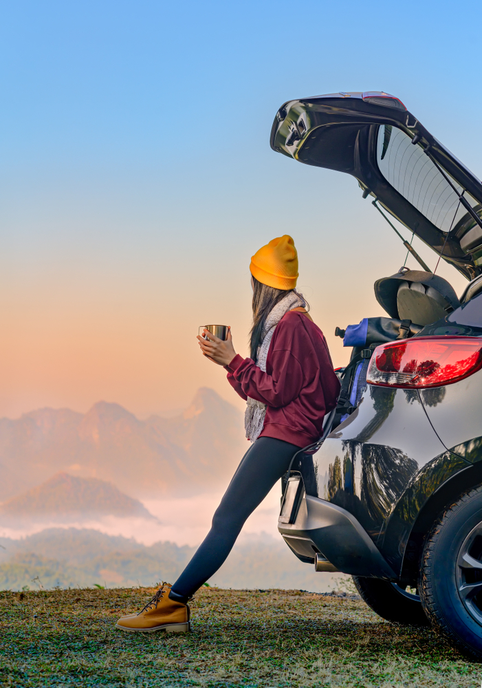 Woman leans against the open hatch of her car holding a warm drink and looking towards the sunset