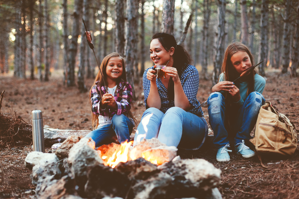 Group of kids with mom sitting by the fire and drinking tea in autumn forest, hike at weekend