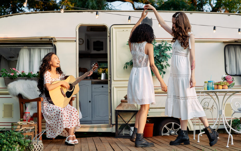 Young happy women have fun together playing guitar and dancing outdoors near their camper van during summer vacation