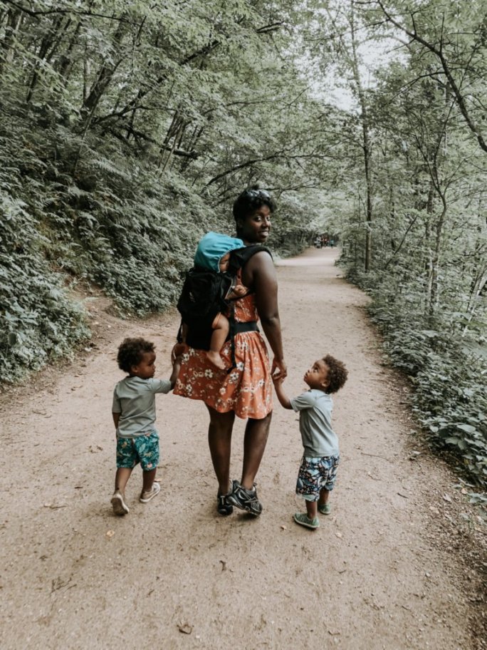 Mother with baby on a back carrier holds hands with two toddlers