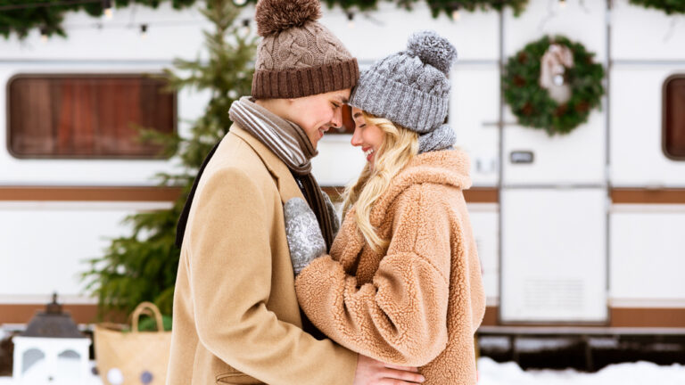 Happy young couple hugging and laughing outdoors in winter, standing near rv trailer motorhome at campsite