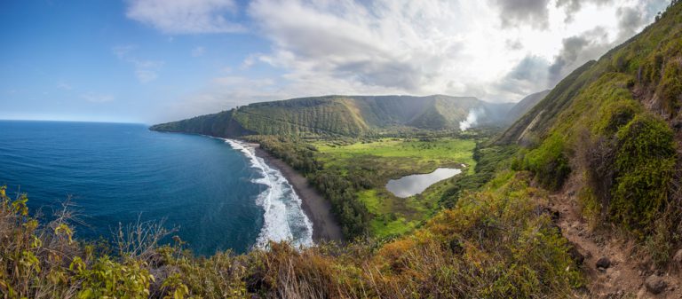 Mountains covered in lush greenery slope to a beach next to the bright blue ocean with a blue sky in the background.