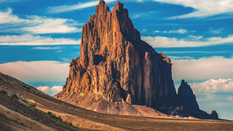 A tall, jagged red-rock butte stands alone in an empty desert under a blue sky.