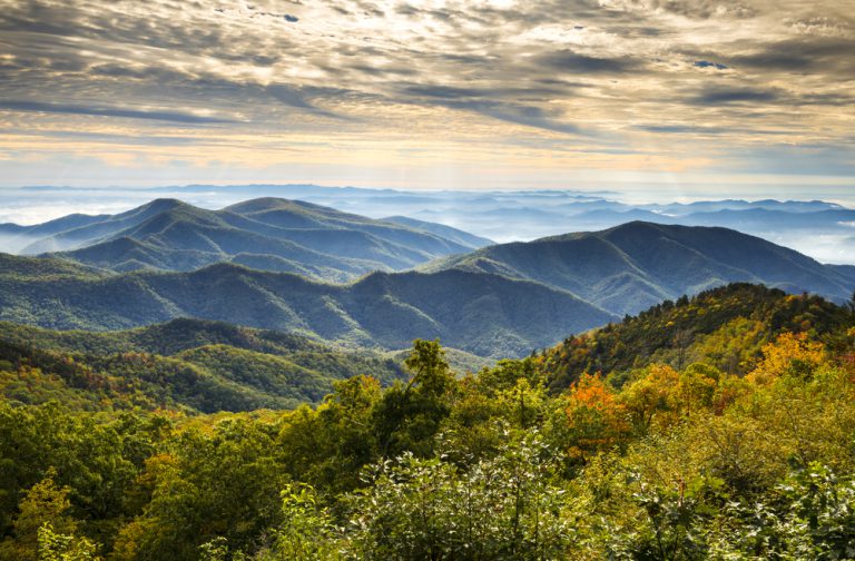 A sweeping mountain range under a cloudy sky with sunlight peeking through and lush fall trees in the foreground.