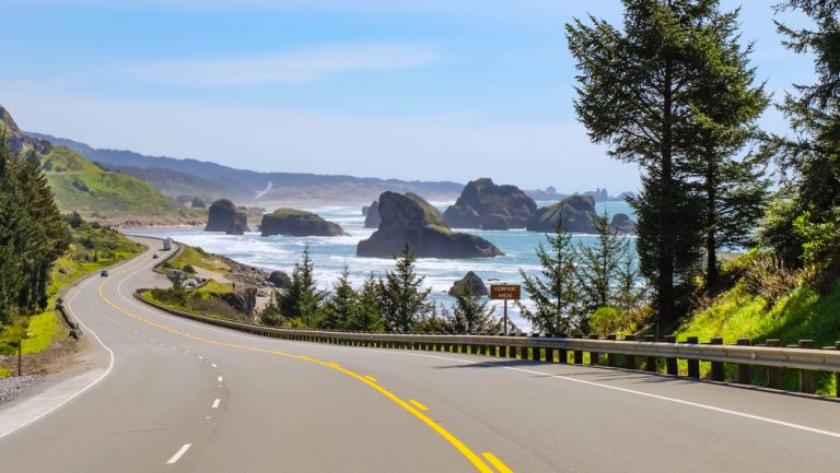 A two-lane road curves to follow a shoreline; rocks sit high in the water under a clear sky.