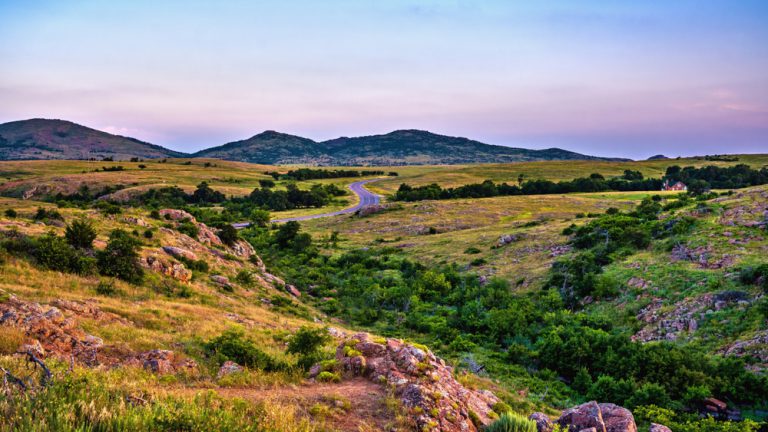 A pink and purple sunset over rolling hills with lines of dense green trees, rocky outcrops, and low hills on the horizon