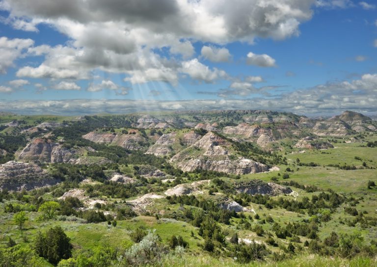 Rays of sunshine break through the clouds above weathered white and red rock bluffs in rolling green hills.