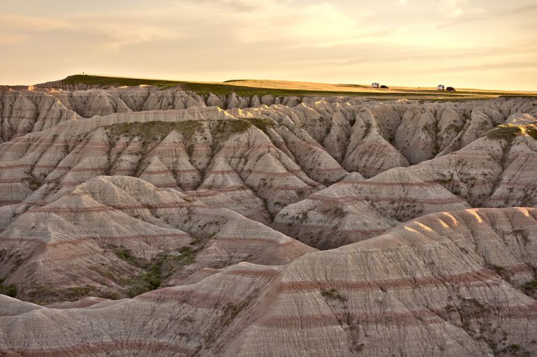 Heavily eroded pink rock formations descend from a plateau, on which two RV's are parked under a yellow, sunset sky.