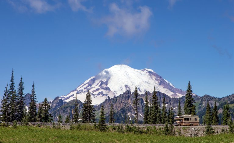 A brown RV drives down a road in front of a snow-peak mountain, evergreen trees dotting the area.