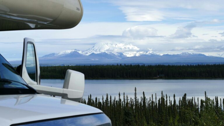 A closeup view of the front of a Class C RV with trees, a shimmering lake and mountains covered in snow in the background