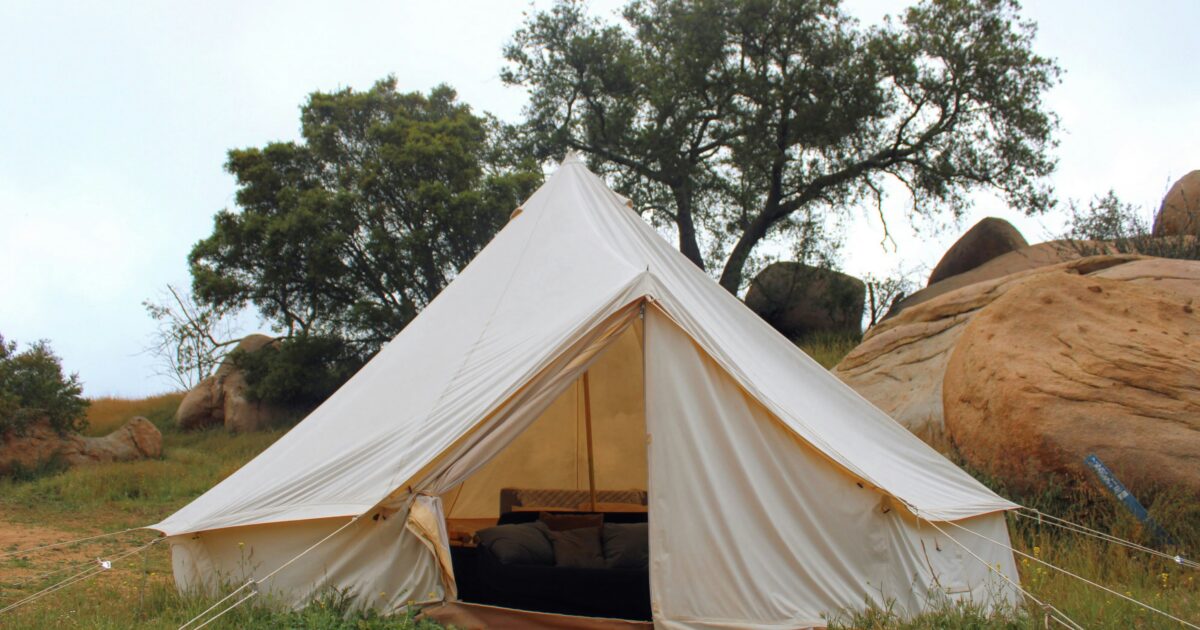 Eggshell-colored safari tent with a view of a bed surrounded by large boulders and large, lush trees and a daytime sky.