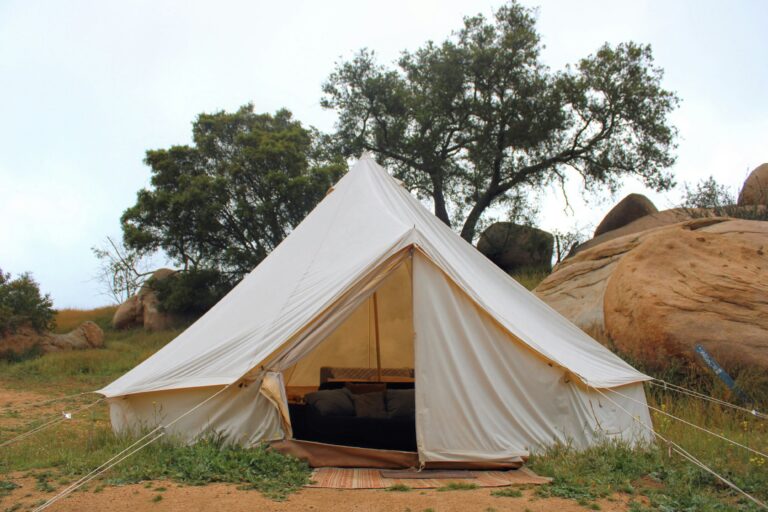 Eggshell-colored safari tent with a view of a bed surrounded by large boulders and large, lush trees and a daytime sky.