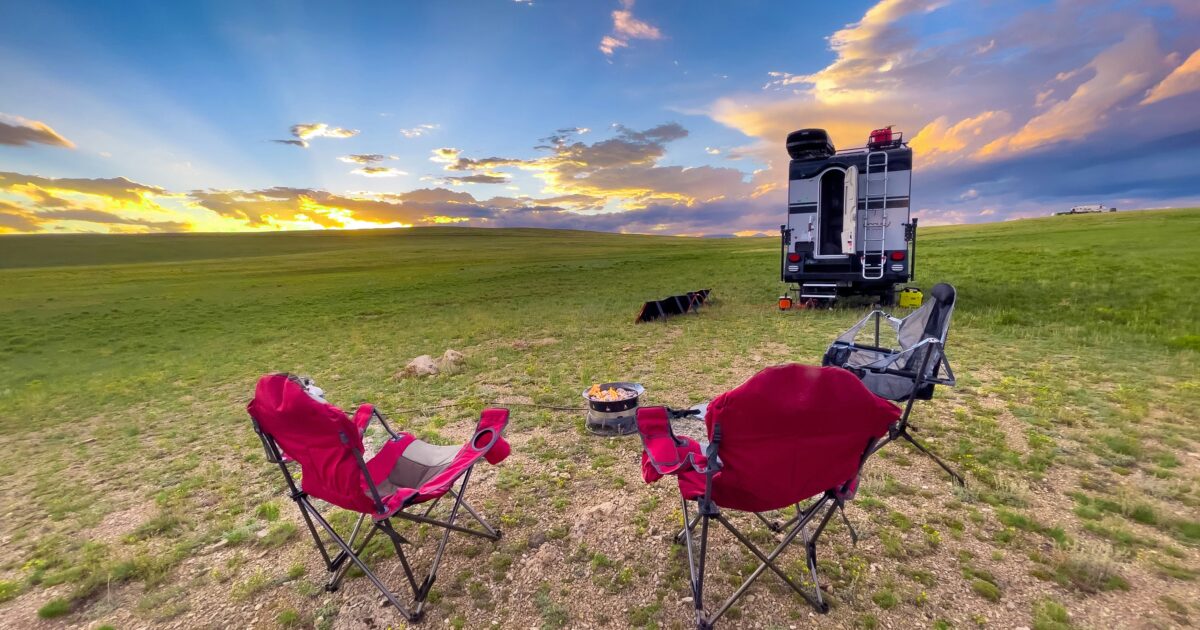 Three folding Muskoka chairs sit in front of the back end of an RV with the sun illuminating a few clouds overhead.