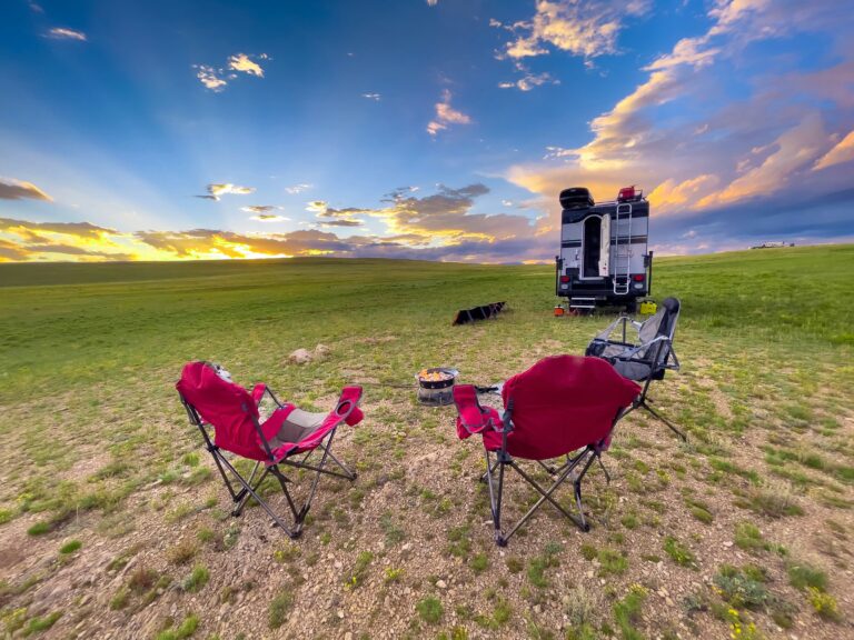 Three folding Muskoka chairs sit in front of the back end of an RV with the sun illuminating a few clouds overhead.