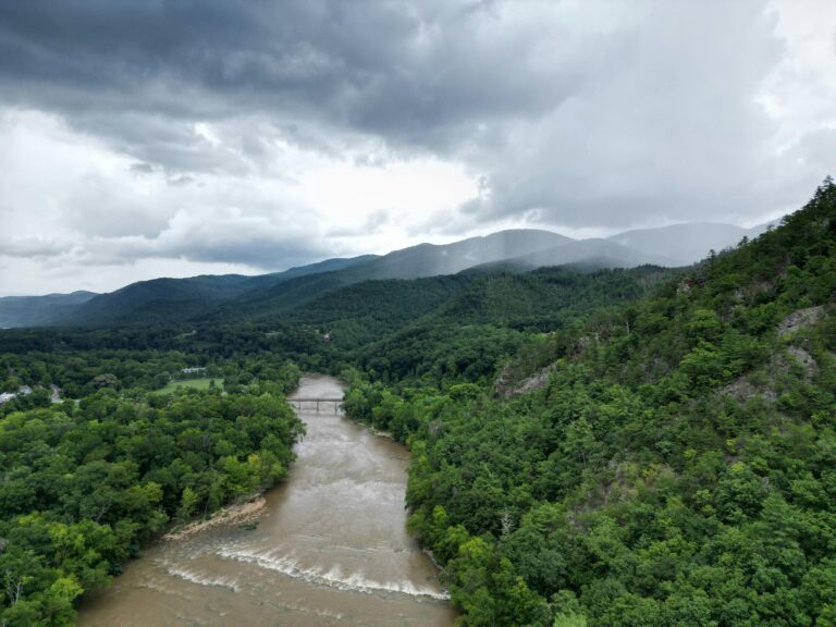 A river flowing through green forests with rolling hills in the background under a cloudy daytime sky.