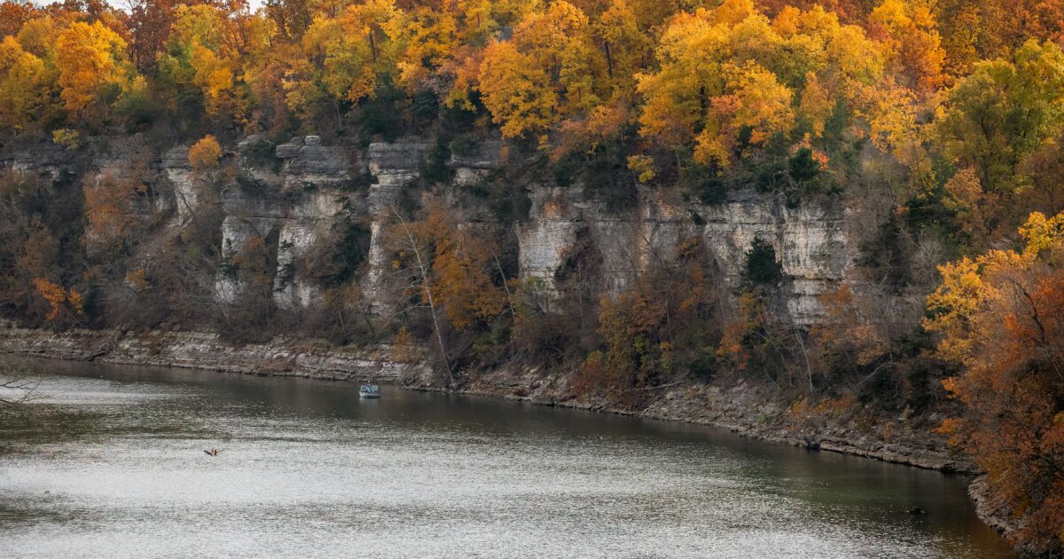 A body of water sits below a cliff topped with trees with browning foliage