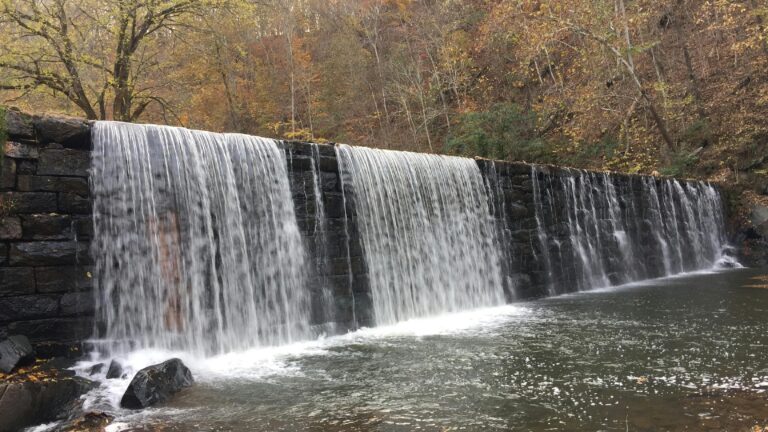A waterfall flowing over a stone wall into a river with browning trees and foliage in the background during the daytime.