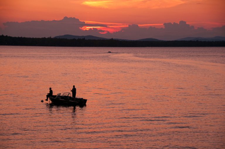 Two people on a boat in the middle of a large, calm lake at sunrise.