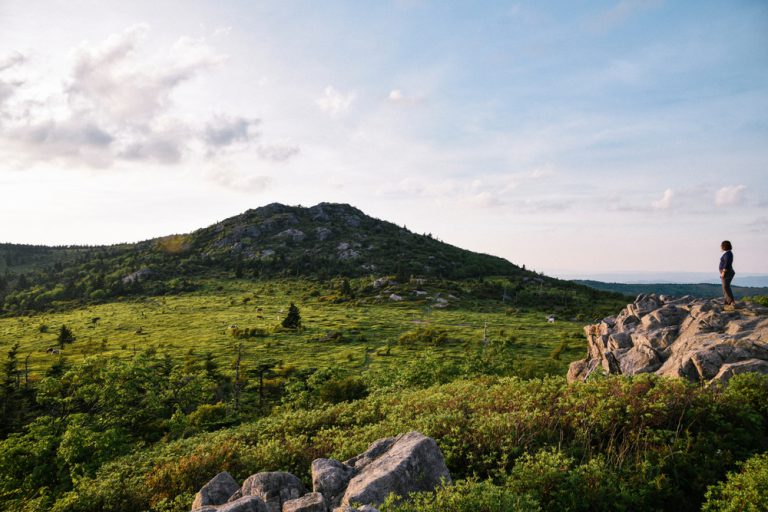 A hiker stands on a rocky outcropping looking out across green fields covering mountains.