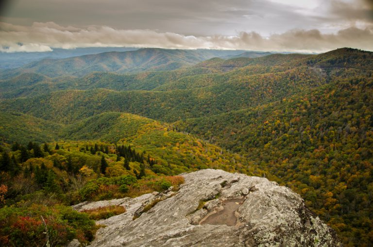 A mountain ridge overlooking a stunning vista of mountains covered in dense forest under a cloud-covered sky.