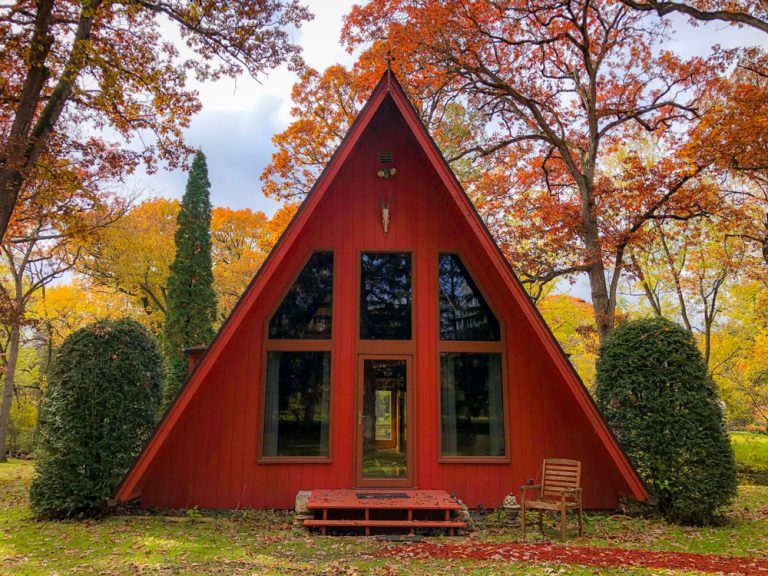 A small, triangular cabin with windows sits in a green field surrounded by short shrubs and tall leafy trees.