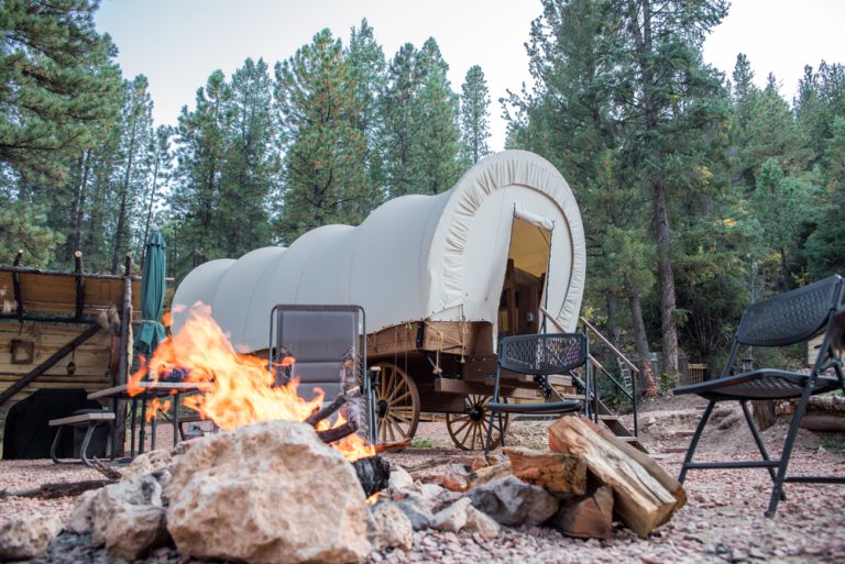 A campfire burns near a Conestoga wagon set in a camping area surrounded by picnic tables, camping chairs, and tall pines