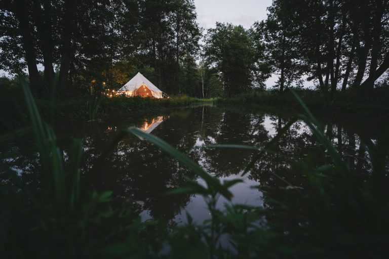 A small white tent with string lights sits along a river surrounded by trees