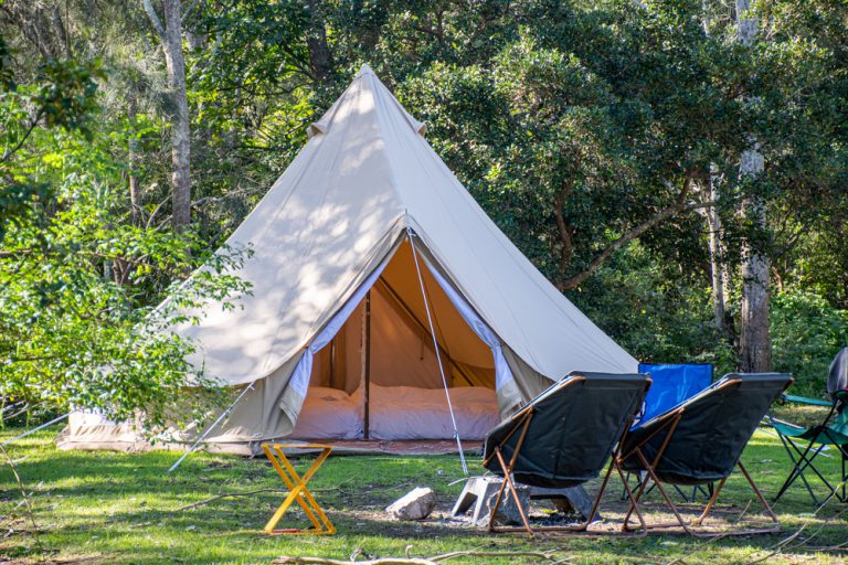 A big white tent with comfortable bedding inside and chairs outside sits on green grass surrounded by trees