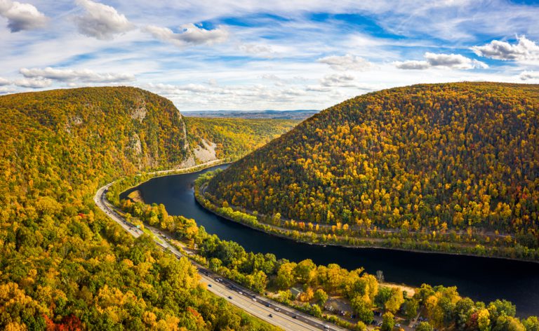 Cars drive on a road that parallels a river, winding between mountain ridges.