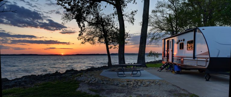 A large RV parked in a concrete camper spot next to a picnic table, set on the shore of a bay of water.