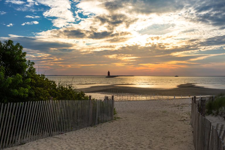 A beach path lined with a short wooden-post fence and green vegetation leads out to a breakwater, the sky lit up with the setting sun and a lighthouse visible in the distance