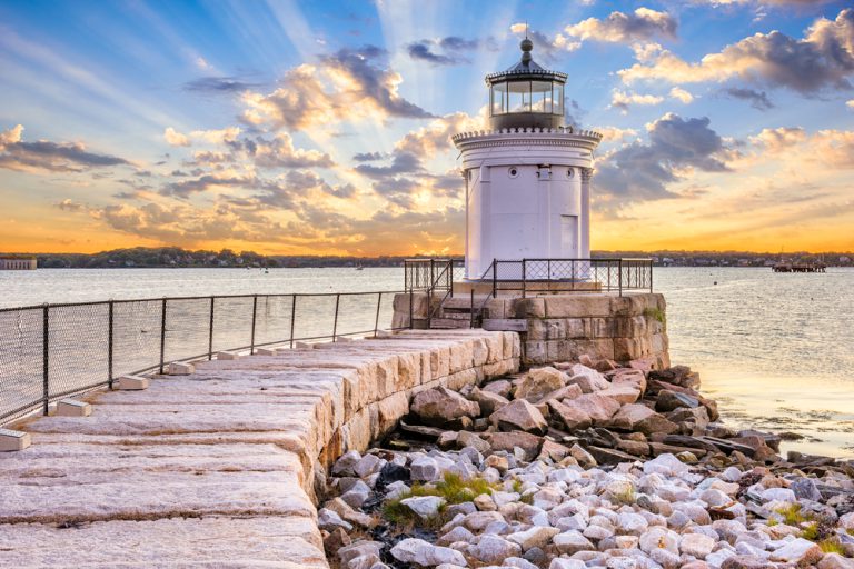 A breakwater on a shore, with a tall lighthouse at its edge. Sunshine peeks through the clouds.