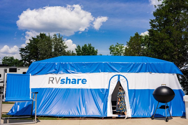 RV with large tent draped over it alongside an oversized camping chair and grill