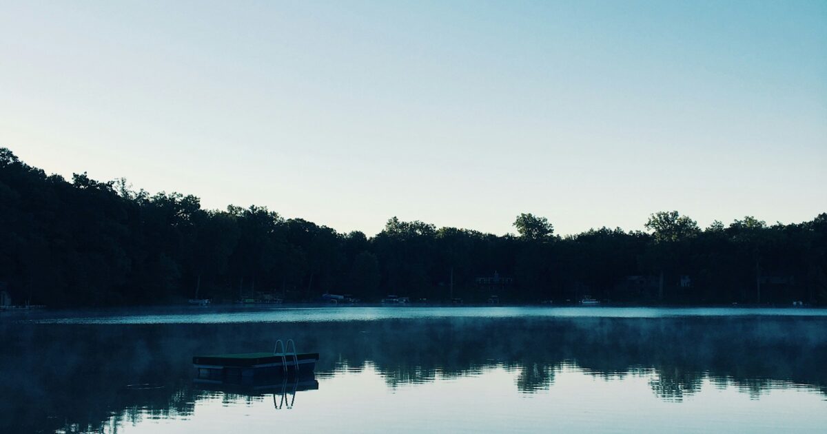 A swimmers' raft sits in the middle of a placid lake in the twilight under a blue sky.