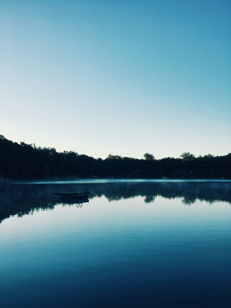 A swimmers' raft sits in the middle of a placid lake in the twilight under a blue sky.