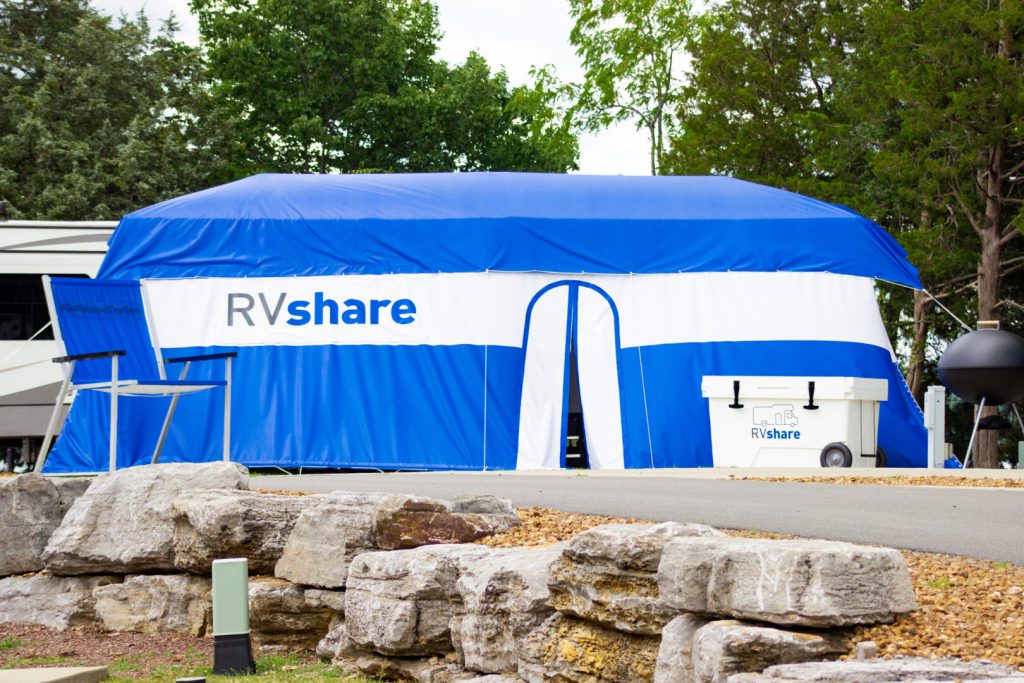 RV with large tent draped over it alongside an oversized camping chair and cooler