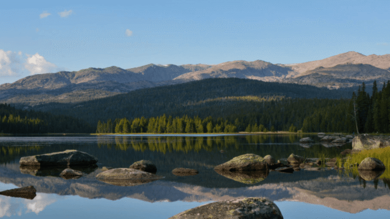 A calm lake reflects a forested bank and mountains under a pale blue sky, great for fishing in Nebraska