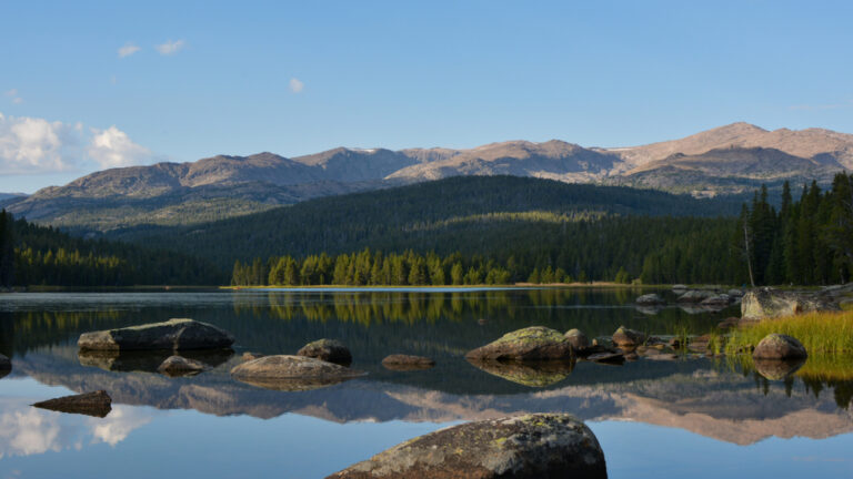 A calm lake reflects a forested bank and tall mountains under a pale blue sky, great for fishing in Nebraska