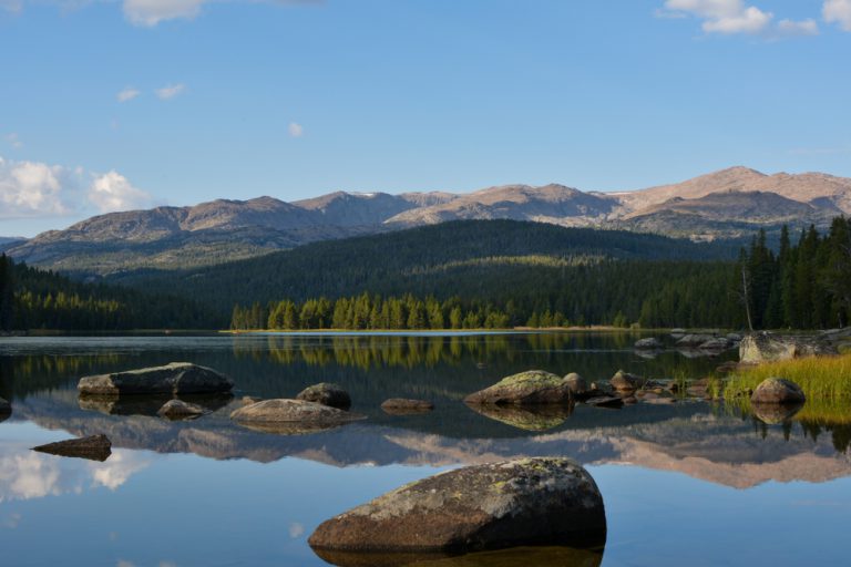 A calm lake reflects a forested bank and tall mountains under a pale blue sky.