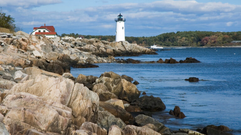 A white lighthouse stands on the rocky shore next to deep blue water, a tug boat sailing in the distance. The shore is surrounded with densely forested banks under a blue sky with white clouds. A great spot for fishing in New Hampshire