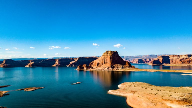 Calm turquoise water in front of weathered red-rock buttes under a blue sky with scattered clouds