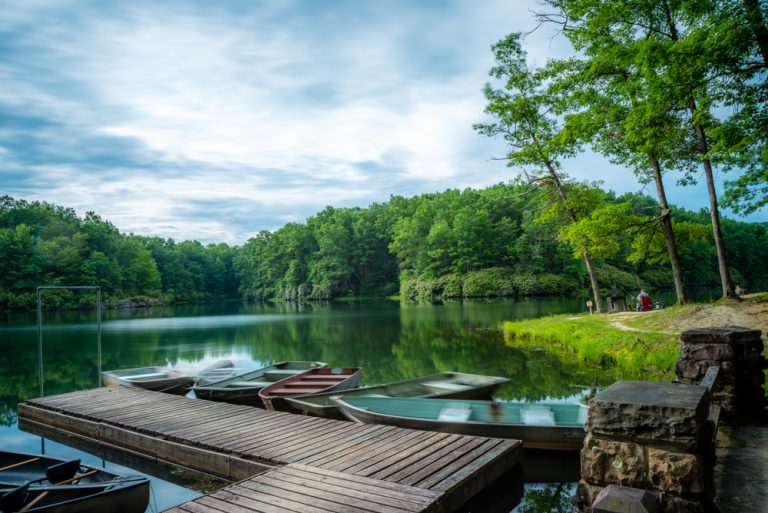 Several small boats at a small wooden dock in a lake surrounded by dense green forest. A person sits, fishing, further down the shore.