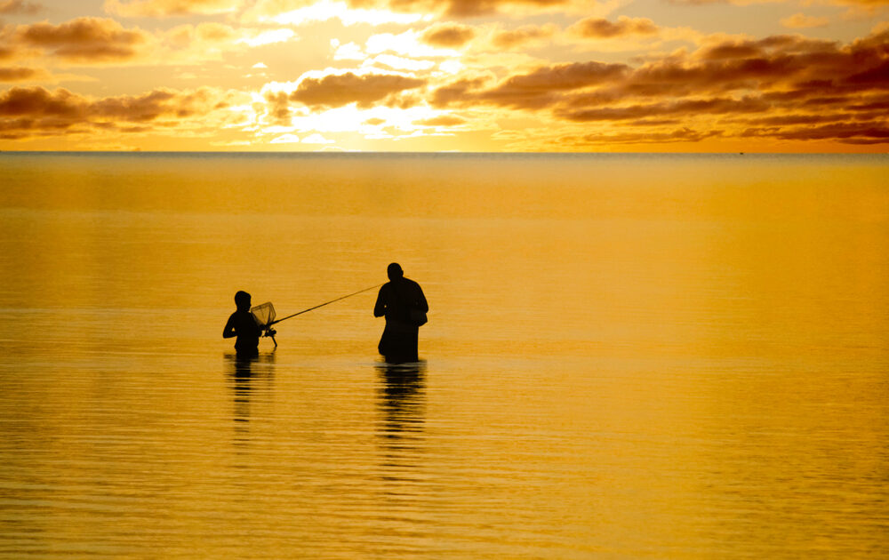A parent and child stand in open ocean water, fishing in North Carolina. The water and sky are orange and yellow with the rising sun.