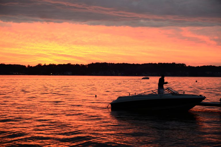 A man in a boat casting a fishing pole, silhouetted by the setting sun.