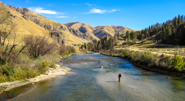 A person stands in the middle of a wide, shallow river surrounded by huge green grass hills.