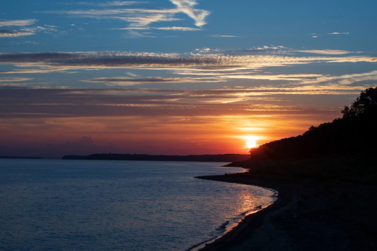 The sun sets behind a shadowed bluff on the bank of a flowing river.