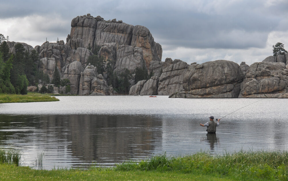 A man stands in the middle of a lake, casting a fishing pole toward deeper waters while fishing in North Dakota. Rocky cliffs make up the other bank.