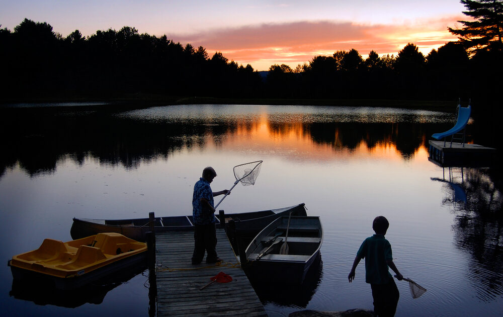 A man and a child standing with nets while fishing in Vermont at a short dock, where a paddle boat and a row boat are docked. The sun rises over trees on the other bank.