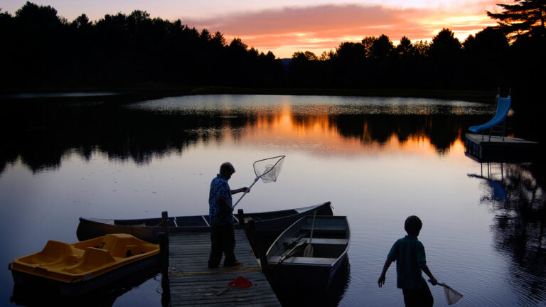 A man and a child standing with nets while fishing in Vermont at a short dock, where a paddle boat and a row boat are docked. The sun rises over trees on the other bank.