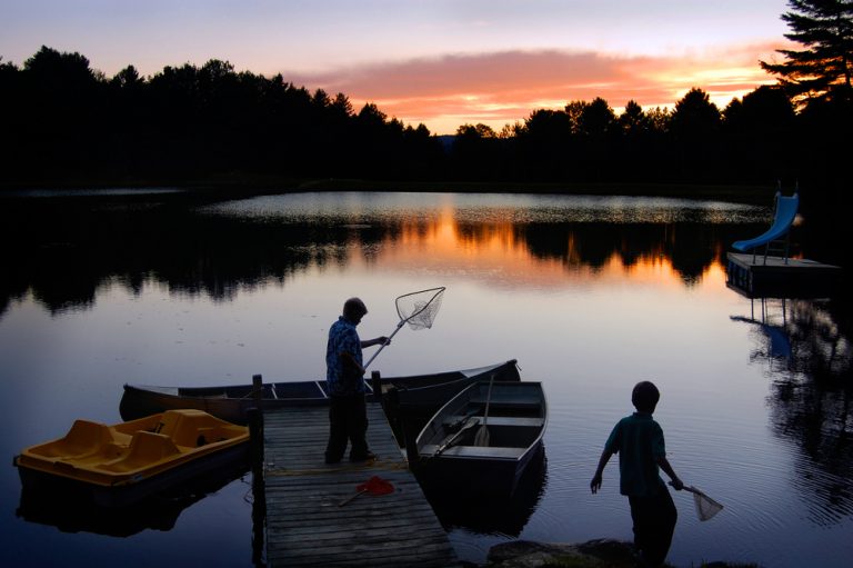 A man and a child standing with nets at a short dock, where a paddle boat and a row boat are docked. The sun rises over trees on the other bank.